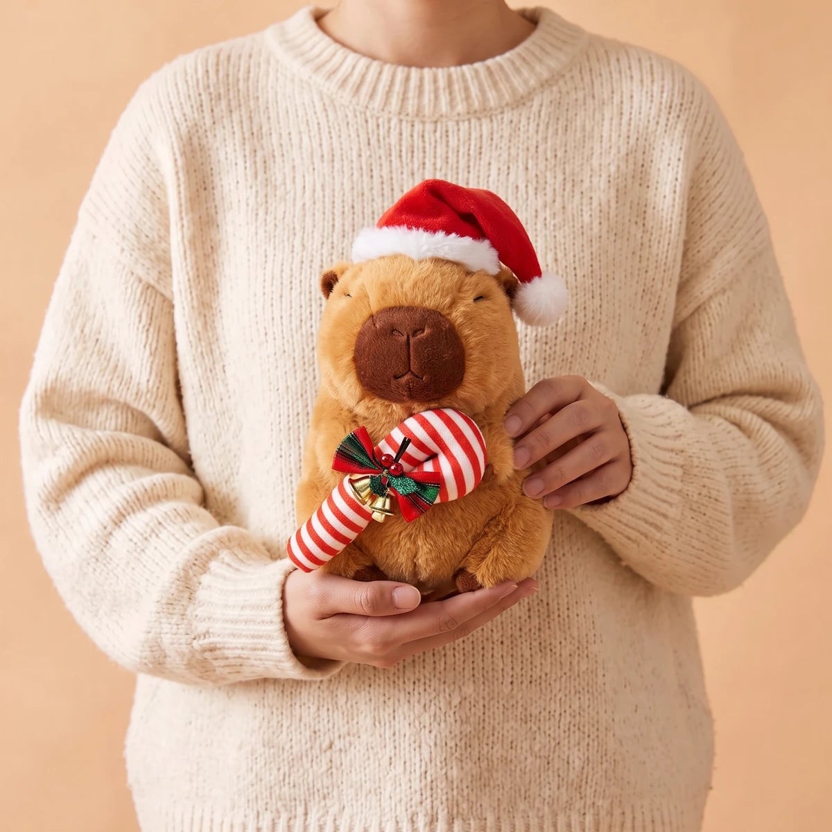 Person in a cream sweater holding the 10 in Christmas capybara plush with a candy cane