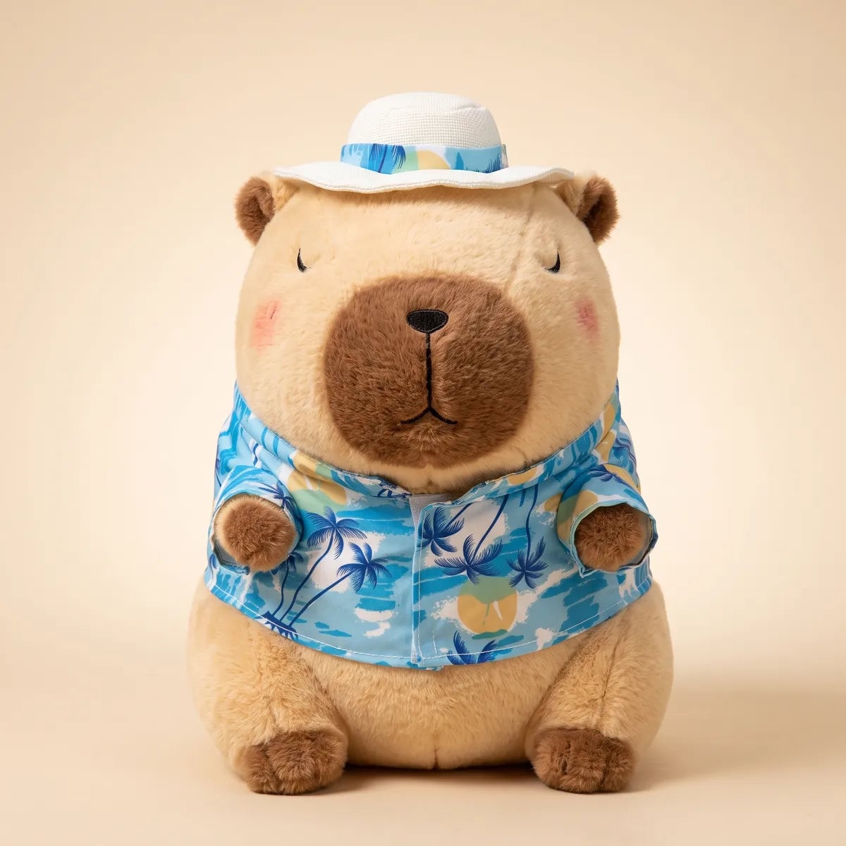 Front view of the brown capybara stuffed animal seated in its blue palm print beach shirt and white sun hat