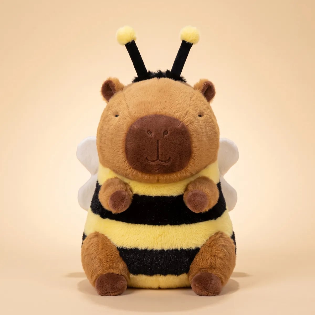 Front view of the capybara bee plush in its yellow and black striped suit with white wings on a beige background