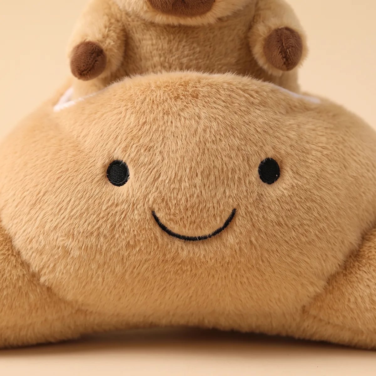 Close up of the croissant face on the capybara croissant plush, two stitched black dot eyes above a curved smile