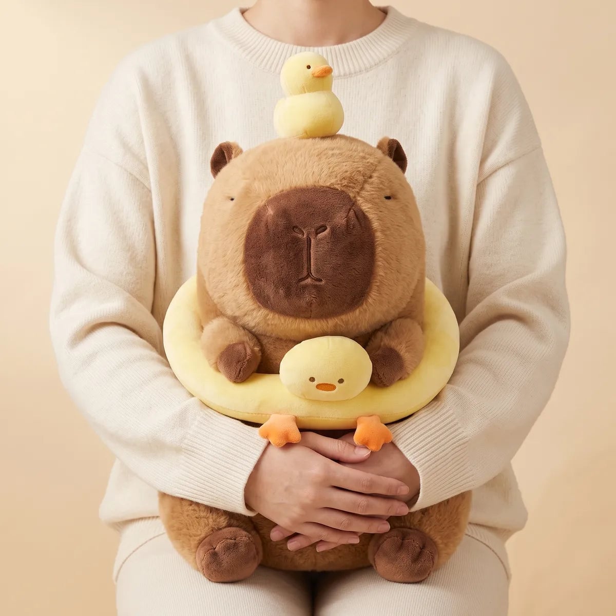 Person holding the capybara duck plush on their lap, forearms passing under the yellow ring and hands clasped at its belly