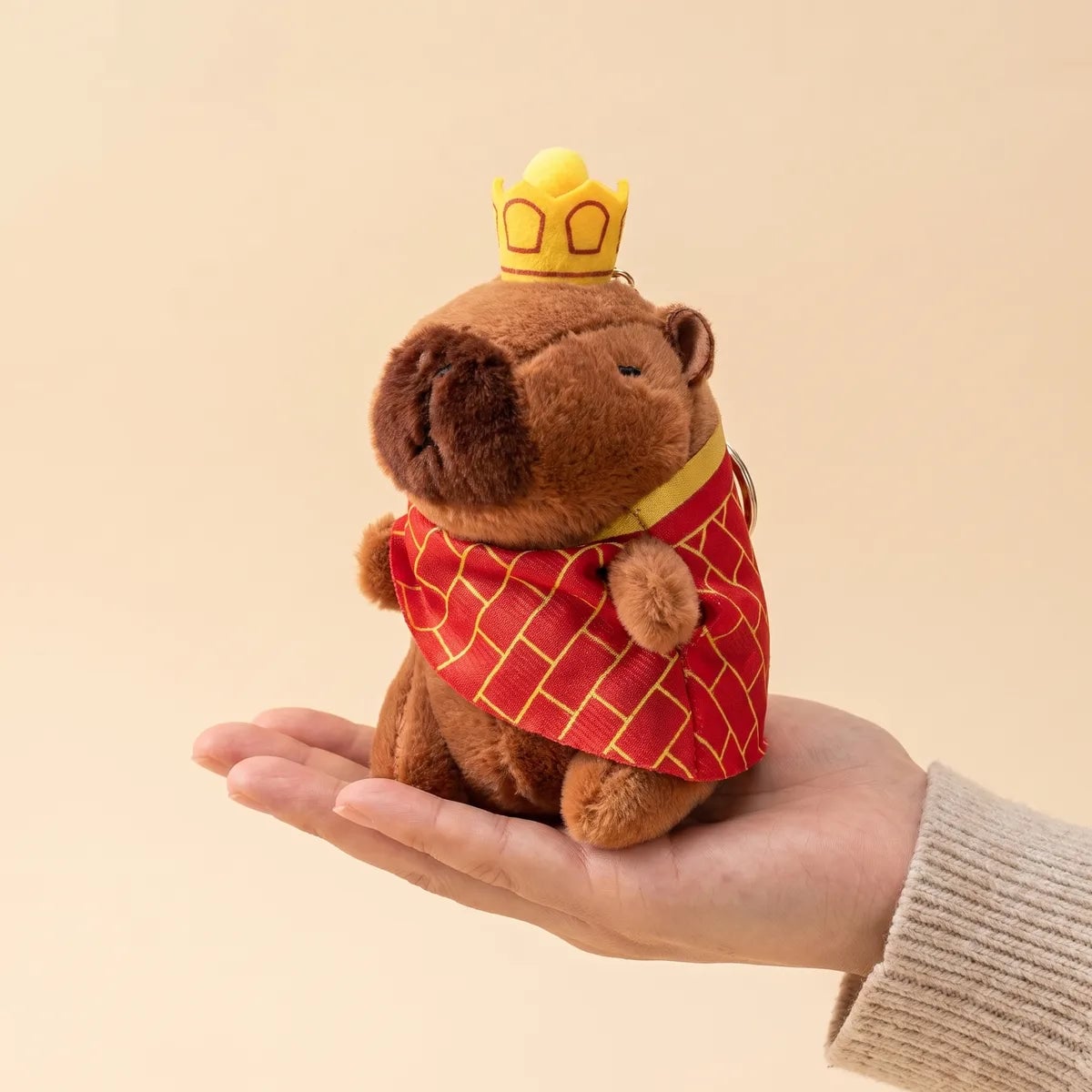 King mini capybara plush standing upright on an open palm with its cape spread over the fingers