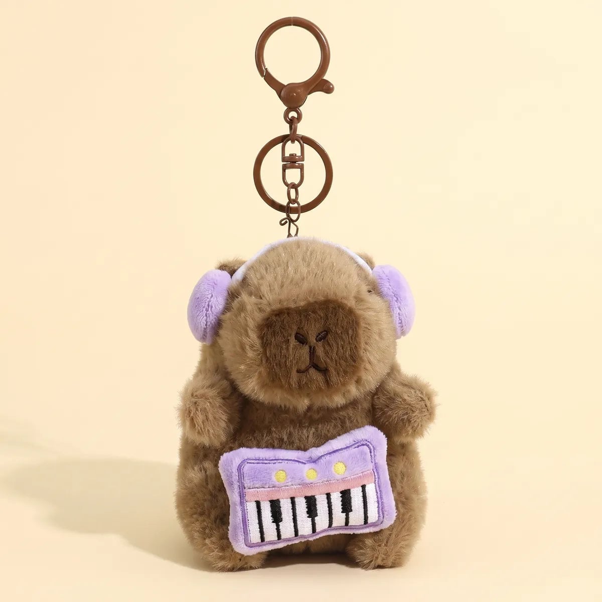 Piano capybara keychain hanging from its bronze clasp with the purple keyboard and lilac headphones facing front
