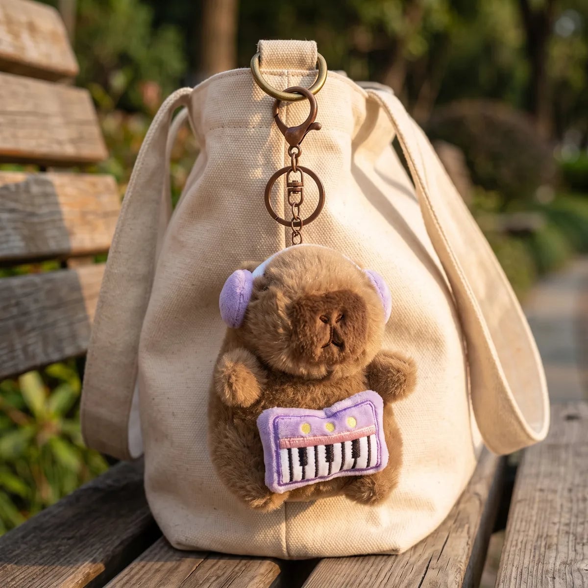 Piano capybara keychain clipped to the handle of a cream canvas tote bag on a park bench