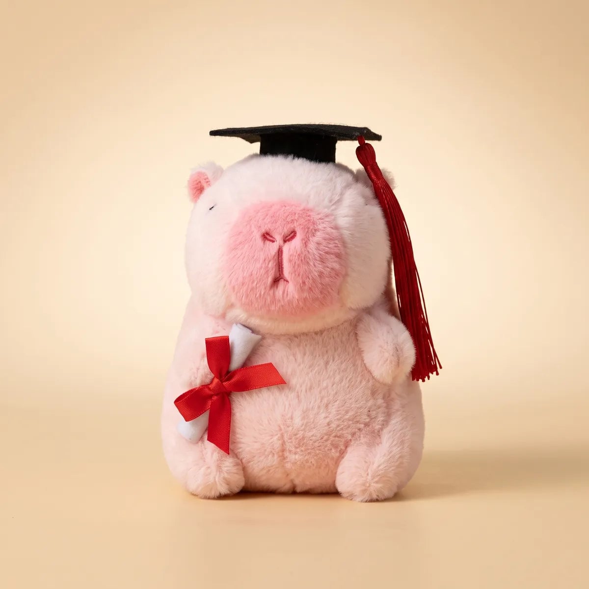 Front view of the pink graduation capybara plush seated on a cream backdrop with its rolled diploma under one arm