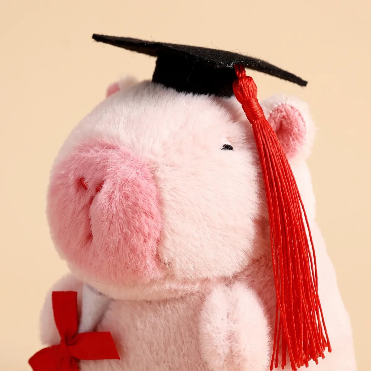 Close-up of the pink graduation capybara plush head showing the black cap, red tassel and stitched eye