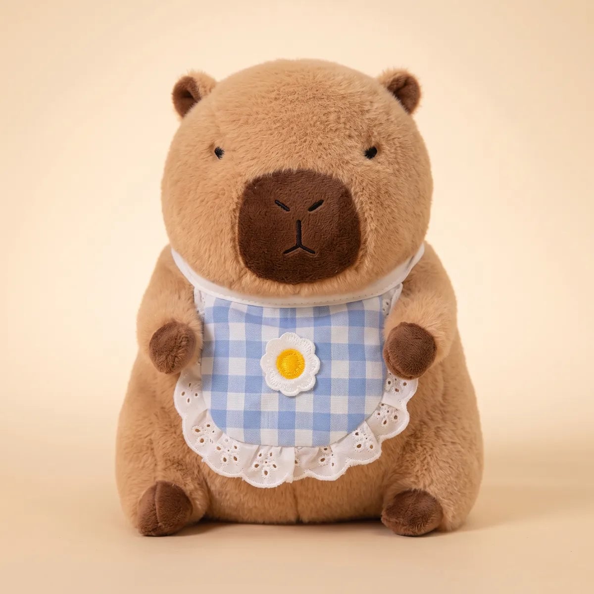 Brown capybara plush seen from the front wearing its blue gingham bib with a daisy