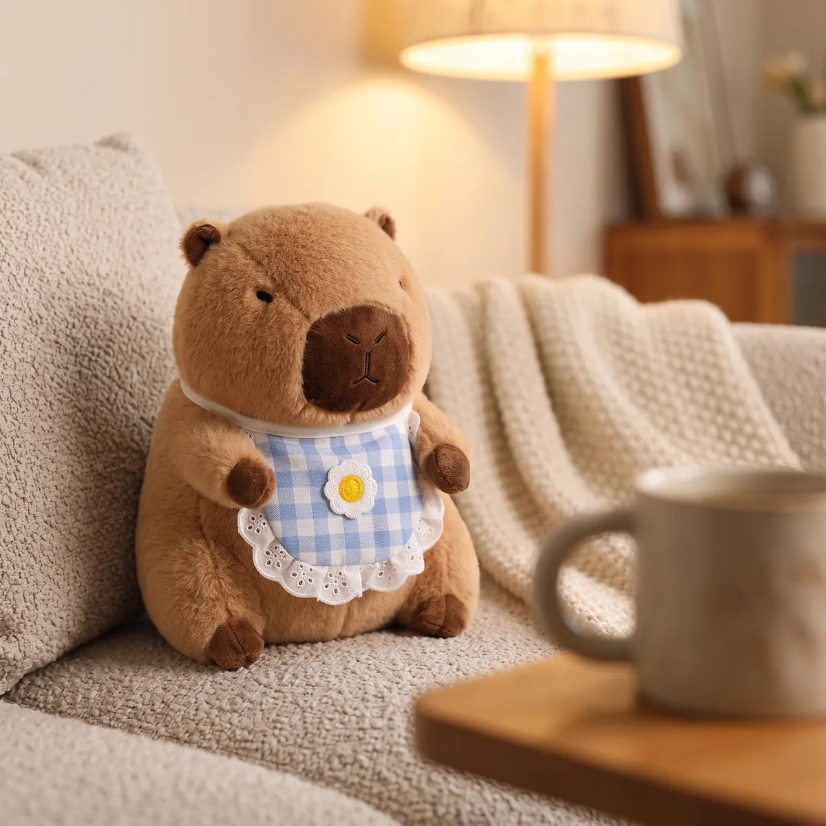 Capybara plush with its gingham bib sitting on a boucle sofa beside a lit lamp