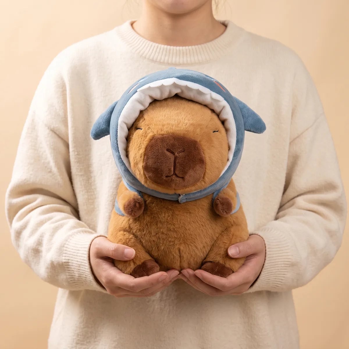 Person holding the shark hat capybara plush against a cream knit sweater