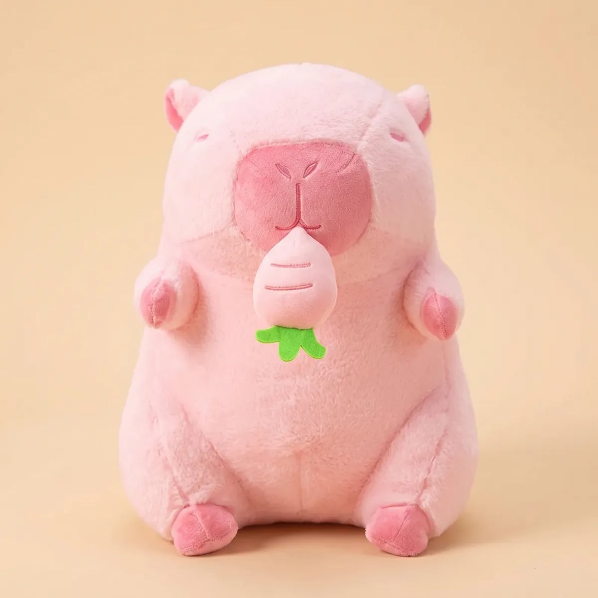 Front view of the 12 in pink capybara plush sitting upright with its turnip against a beige backdrop