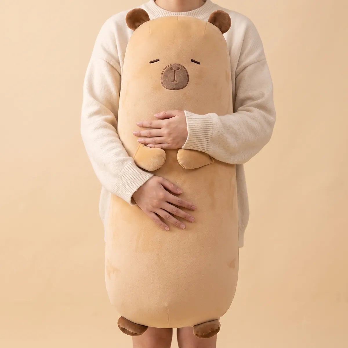 Big capybara stuffed animal plush held upright against an adult, the sand colored body reaching from mid thigh to shoulder