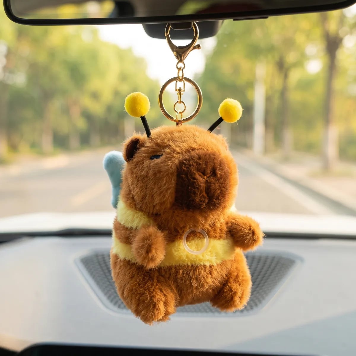 Capybara bee keychain plush hanging from a car rear view mirror with a tree lined road in the background