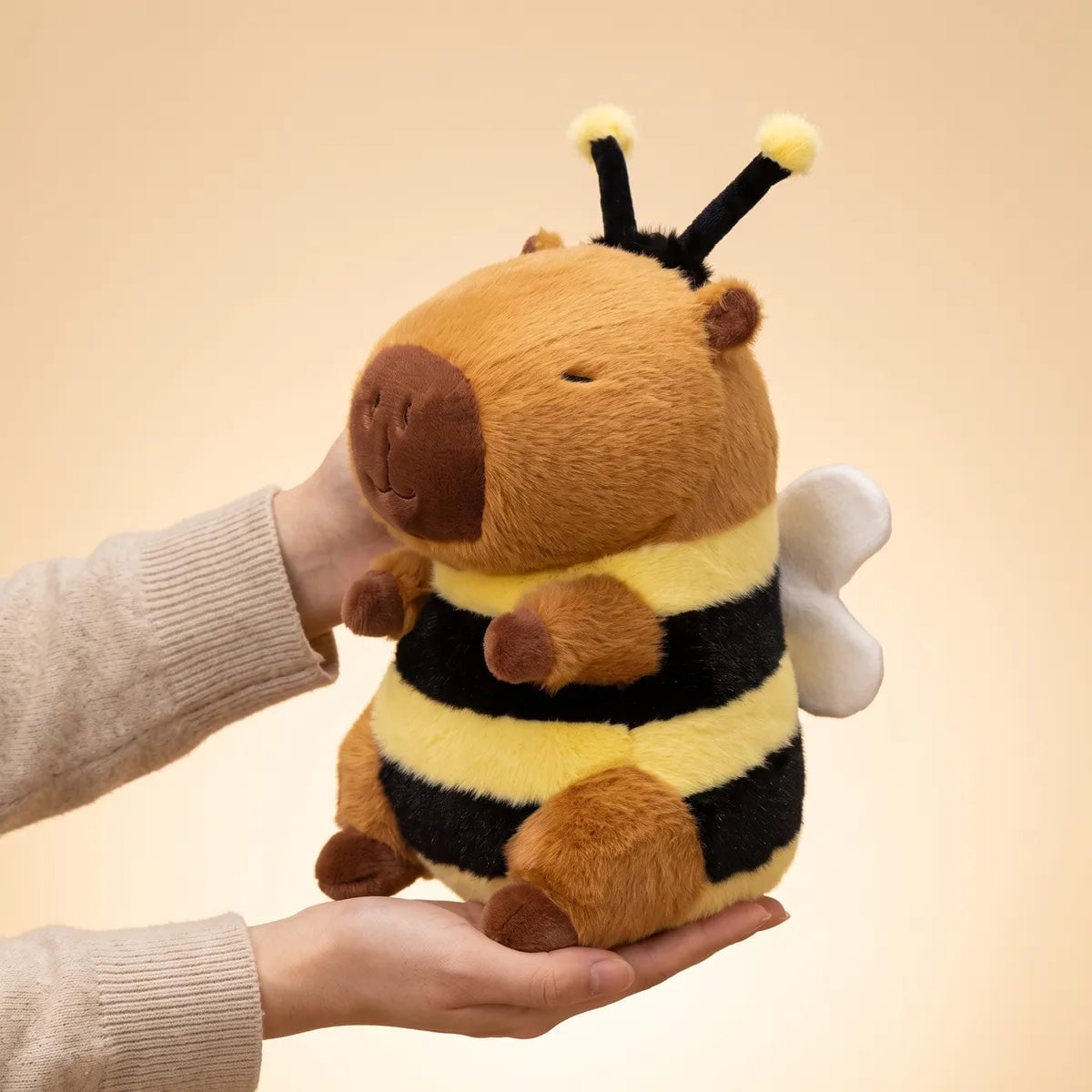 Capybara bee plush held in two hands, seen from the side with one white wing showing behind the striped body