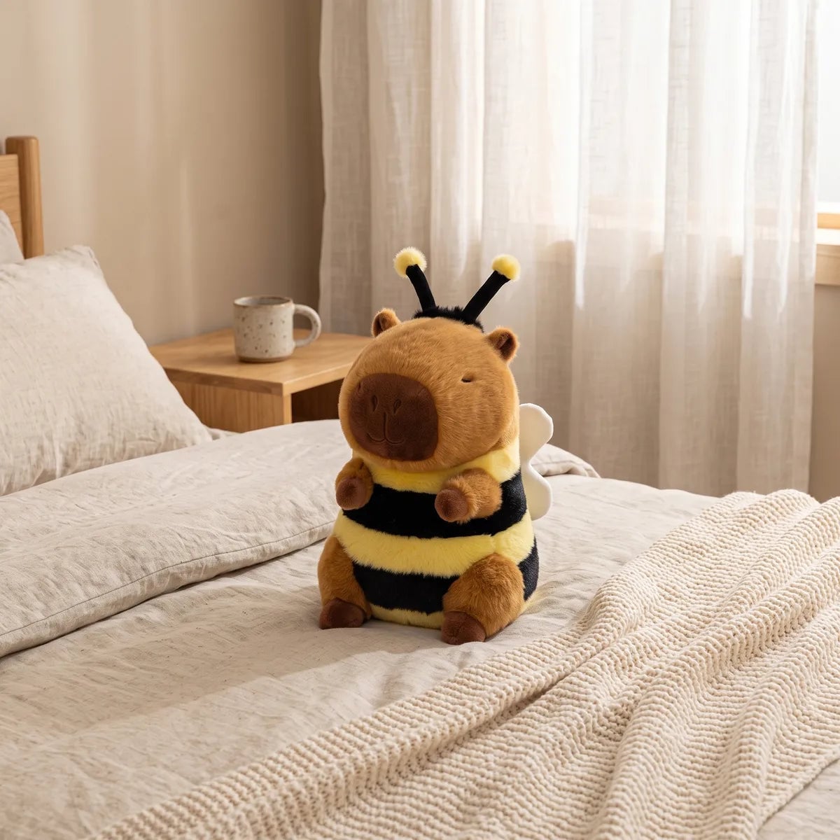 Capybara bee plush upright on the bedding with its white wings clear of the pillow and a mug behind it
