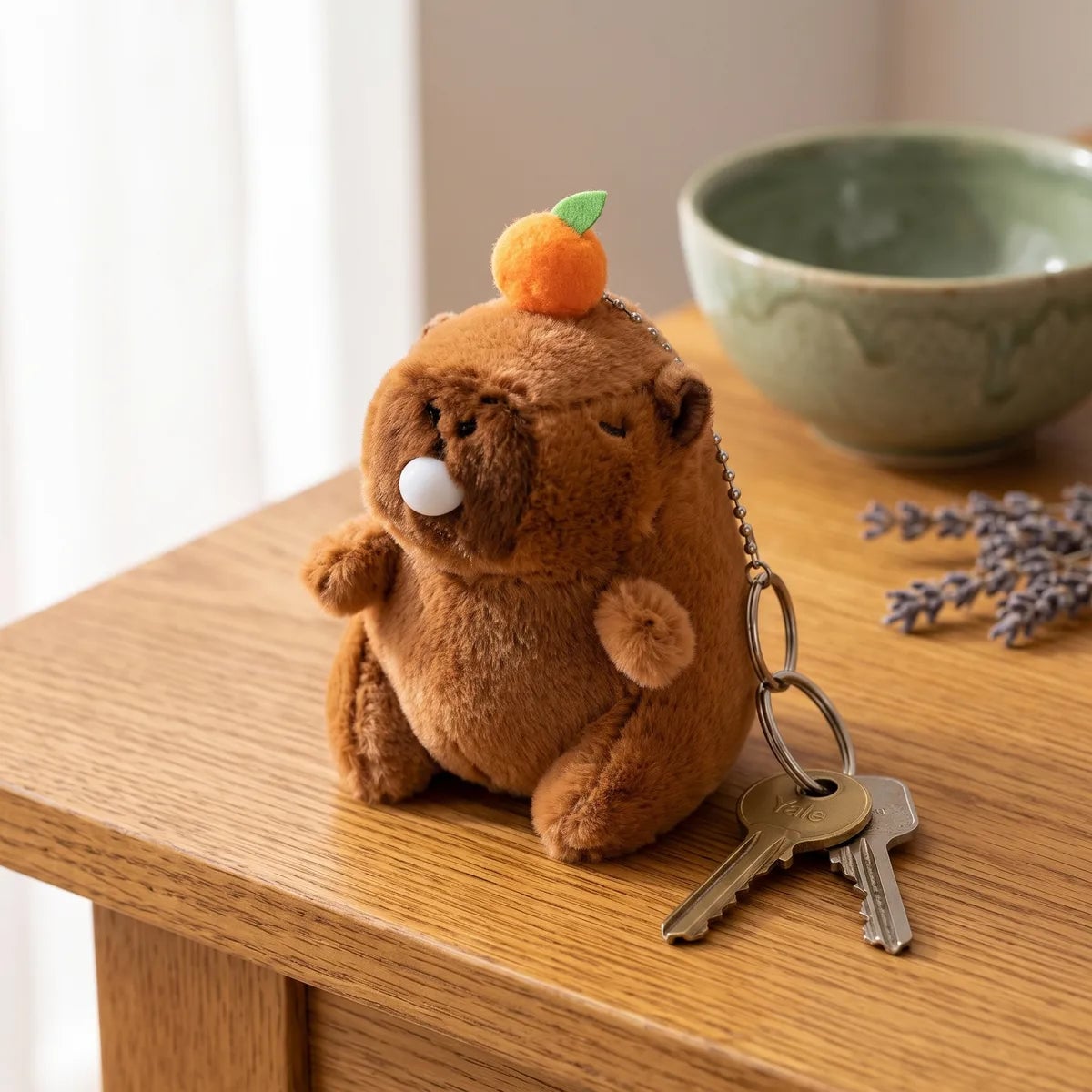 Capybara blowing bubble keychain plush resting on a wood table beside a brass key and a silver key on its split ring
