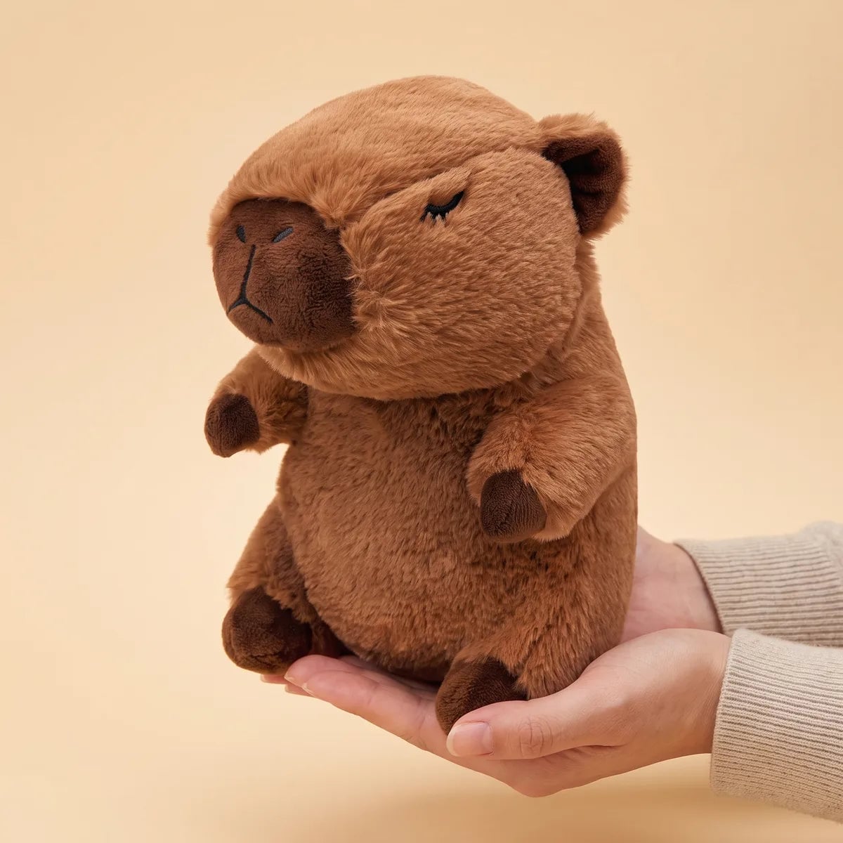 Capybara breathing plush resting across two cupped hands, showing its 13 inch seated height against an adult palm.