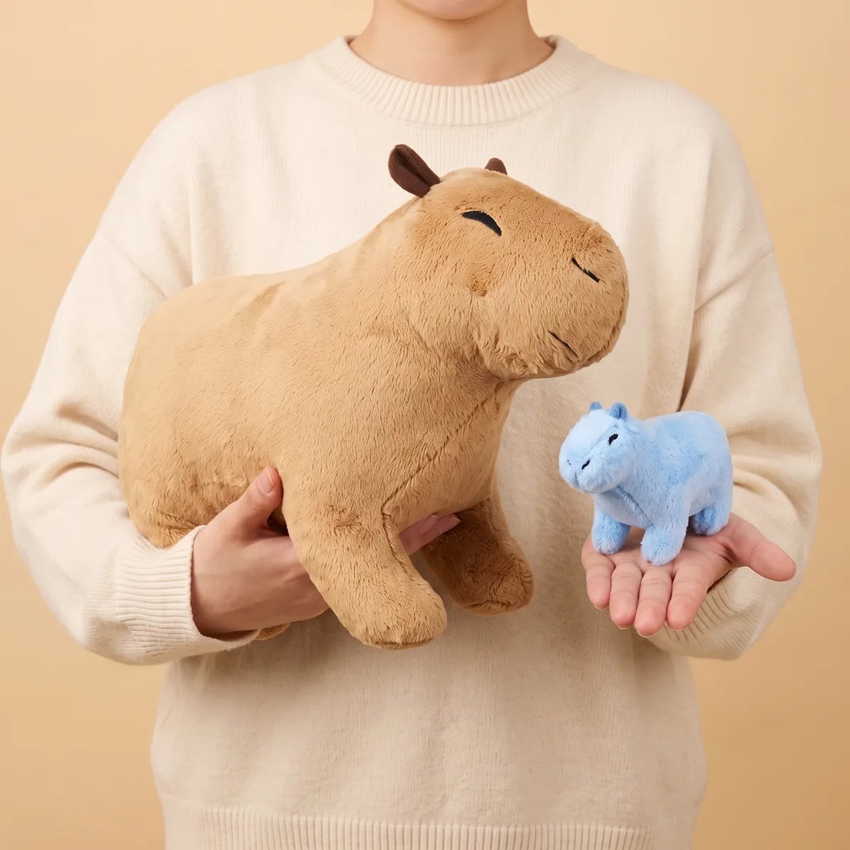 Person holding the tan mother capybara plush in both hands with a blue baby capybara resting on one palm