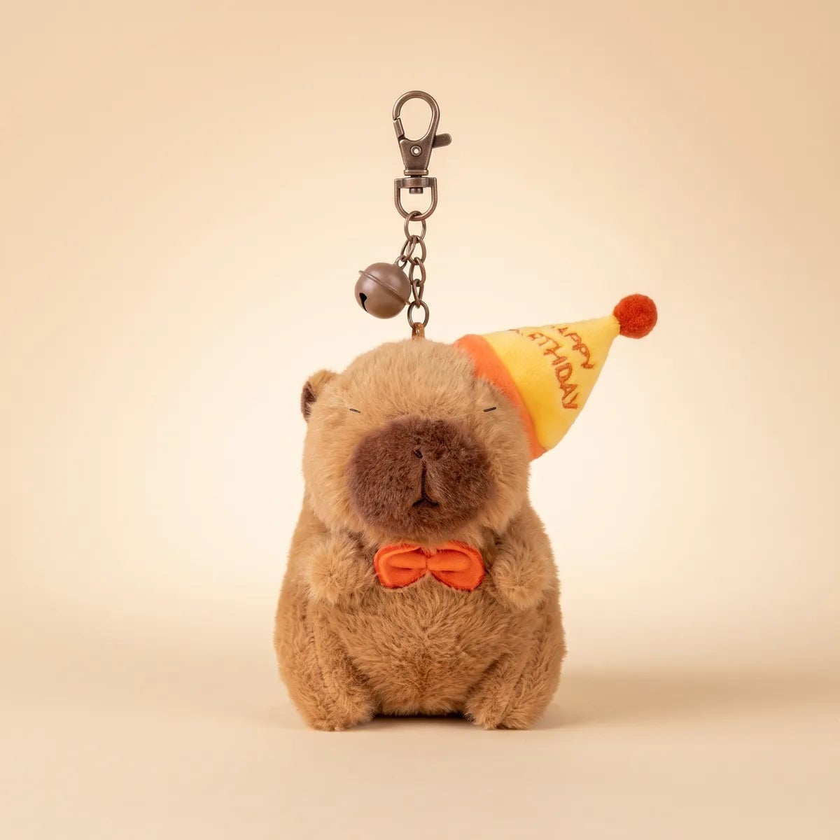 Front view of the birthday capybara keychain hanging from its chain, bell and lobster clasp on a beige backdrop