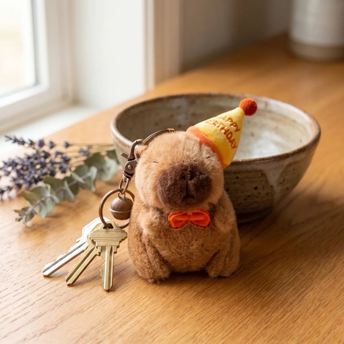 Birthday capybara keychain attached to three keys on a wooden table beside a ceramic bowl