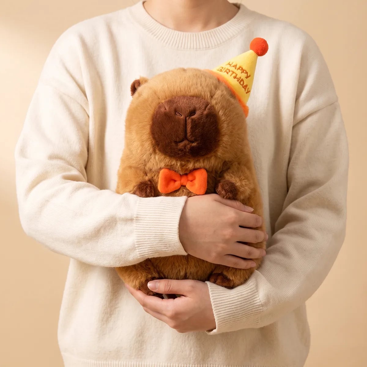 Person in a cream sweater carrying the 13 in happy birthday capybara plush in both arms