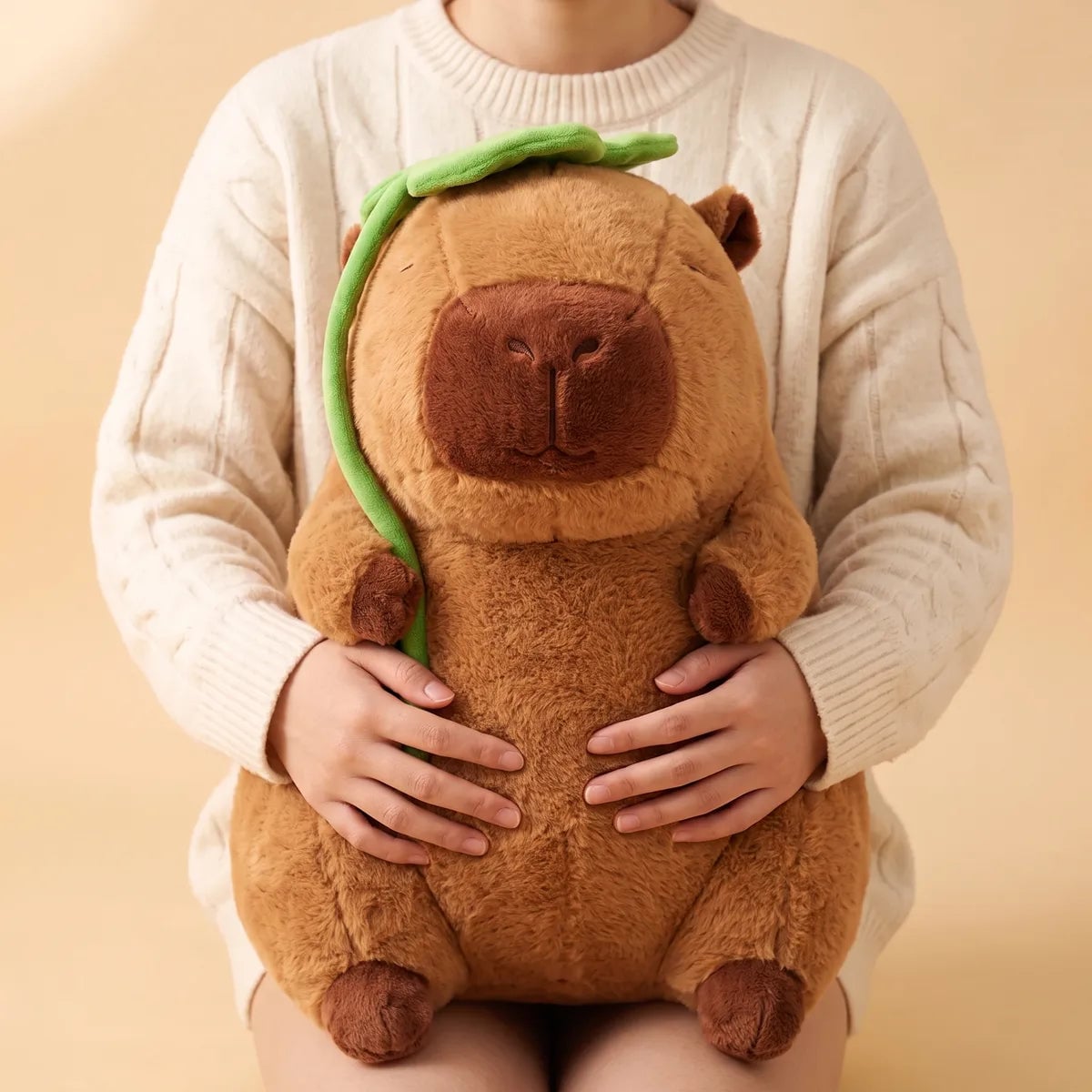 Adult in a cable knit sweater holding the jumbo capybara plush upright on their lap with both hands.