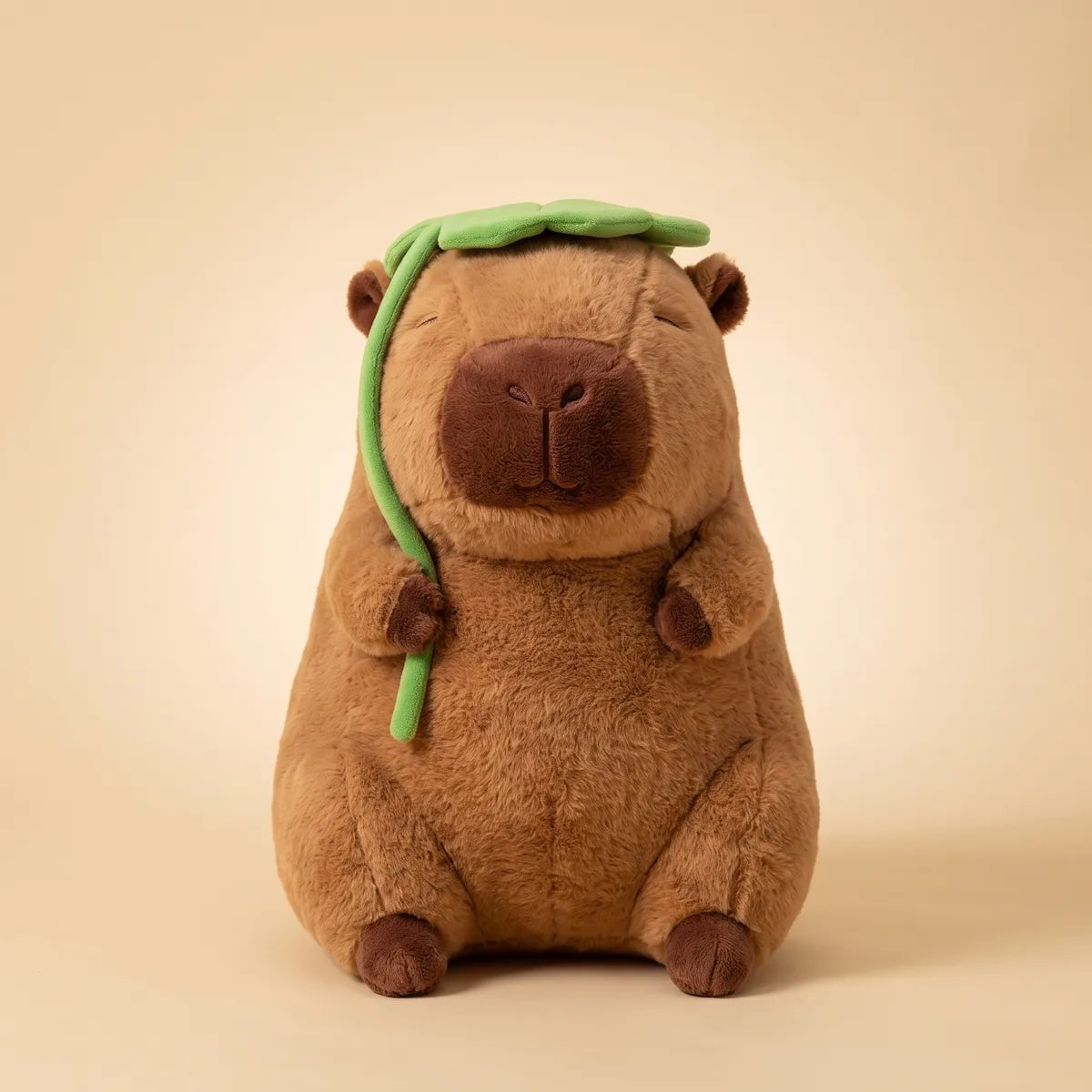 Jumbo capybara plush seated front on against a cream studio backdrop with the green leaf over its head.