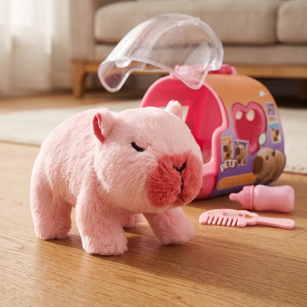 Pink walking capybara toy standing on a wood floor beside the open carry case, a comb and a bottle