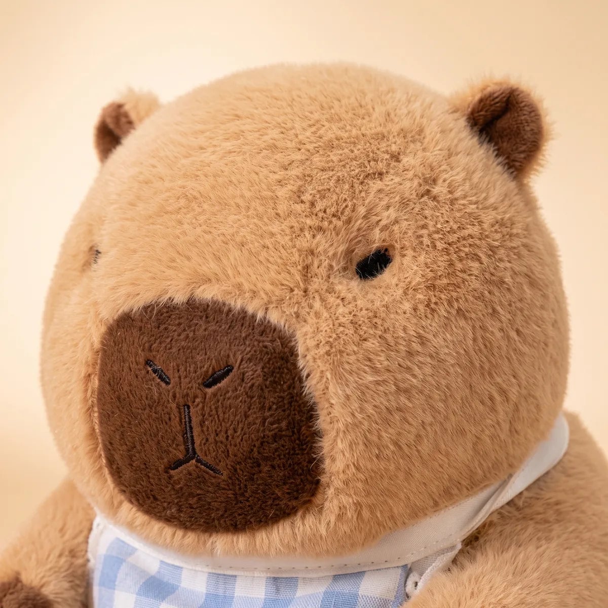 Close up of the capybara plush face and its embroidered muzzle above the gingham bib