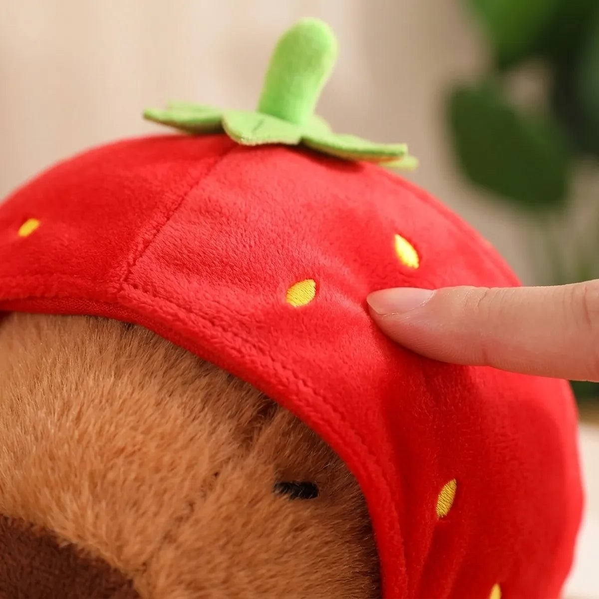 Fingertip pressing the soft red hood fabric of the strawberry capybara plush and its embroidered seeds