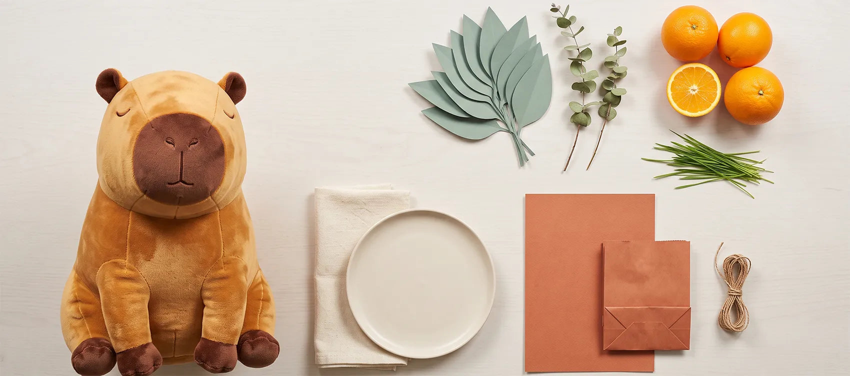 Flat lay of capybara party decorations: capybara plush, paper leaves, eucalyptus, oranges, a plate, kraft card and twine